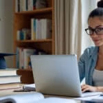 Ein konzentrierter Graduierungsstudent sitzt an einem modernen Schreibtisch mit Laptop, Tablet und Lehrbüchern in einem hell beleuchteten Heimstudierzimmer mit Bücherregal und Graduierungshut im unscharfen Hintergrund.
