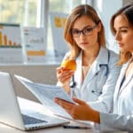 Eine professionelle Ernährungsberaterin im weißen Kittel analysiert einen Ernährungsplan an ihrem modernen Schreibtisch mit Laptop, während sie einen roten Apfel hält, umgeben von Ernährungstabellen und medizinischen Fachbüchern in einem hell beleuchteten Praxisraum.