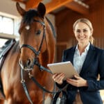 Eine professionelle Reitlehrerin in klassischer Reitbekleidung steht selbstbewusst neben einem gepflegten braunen Pferd in einem modernen, lichtdurchfluteten Holzstall während sie Reitutensilien und Lehrmaterial in den Händen hält.