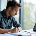 Ein konzentrierter junger Student in legerer Businesskleidung arbeitet an einem modernen Schreibtisch mit Laptop, Lehrbüchern und digitalen Lernmaterialien in einem natürlich beleuchteten Studienumfeld.