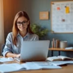 Eine junge Geschäftsfrau im Smart-Casual-Outfit sitzt an einem modern eingerichteten Home-Office-Schreibtisch mit Laptop, Studienmaterialien und Kaffeetasse, während warmes Tageslicht durch ein nahegelegenes Fenster fällt und ein Wandkalender sowie eine Pinnwand im Hintergrund ihre organisierte Arbeitsweise unterstreichen.